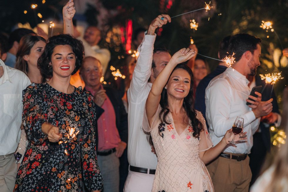 Party guests celebrating at a festive outdoor event with sparklers under glowing string lights.