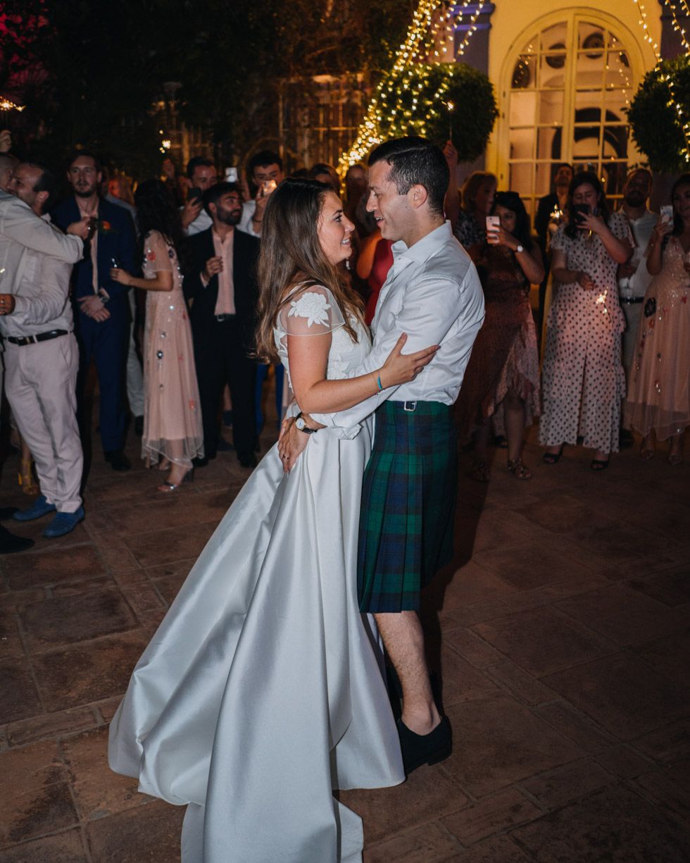 A couple dancing at a wedding celebration with the bride in white and groom wearing a traditional Scottish kilt.