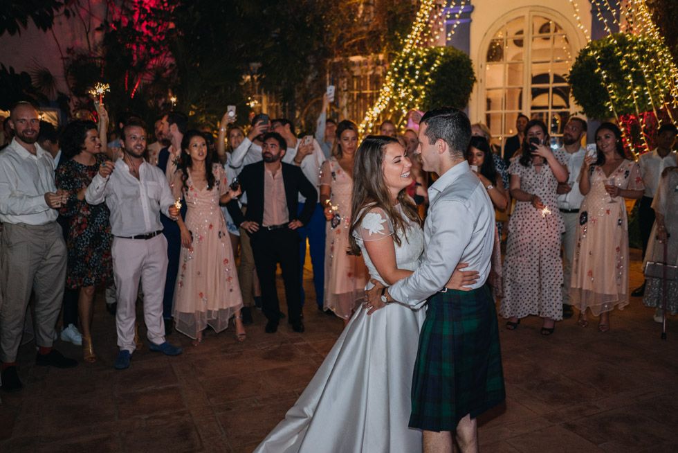 A couple shares their first dance at a nighttime wedding reception while guests watch from the courtyard illuminated by twinkle lights.