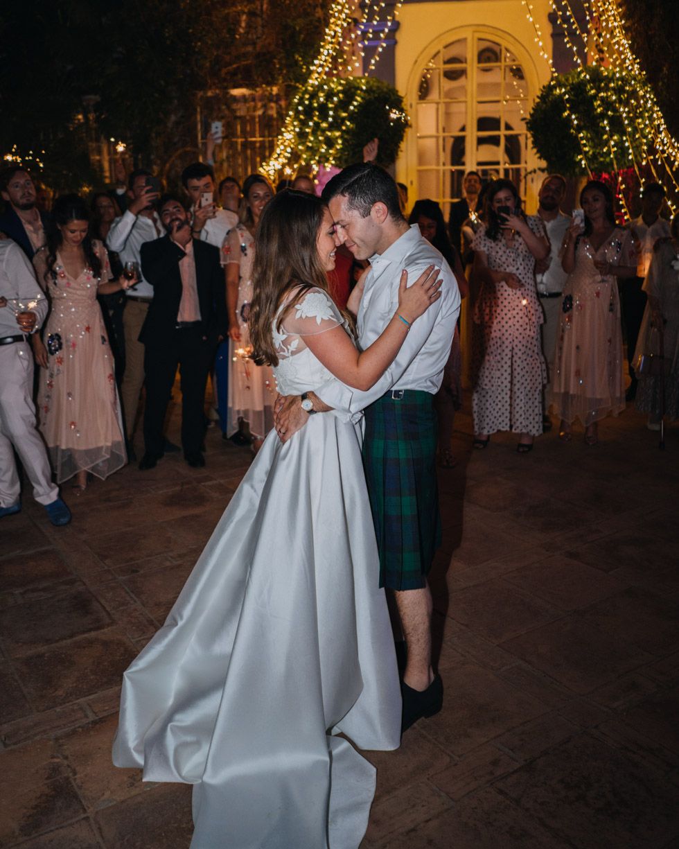 Newlywed couple sharing their first dance at night under illuminated trees while wedding guests watch them celebrate.