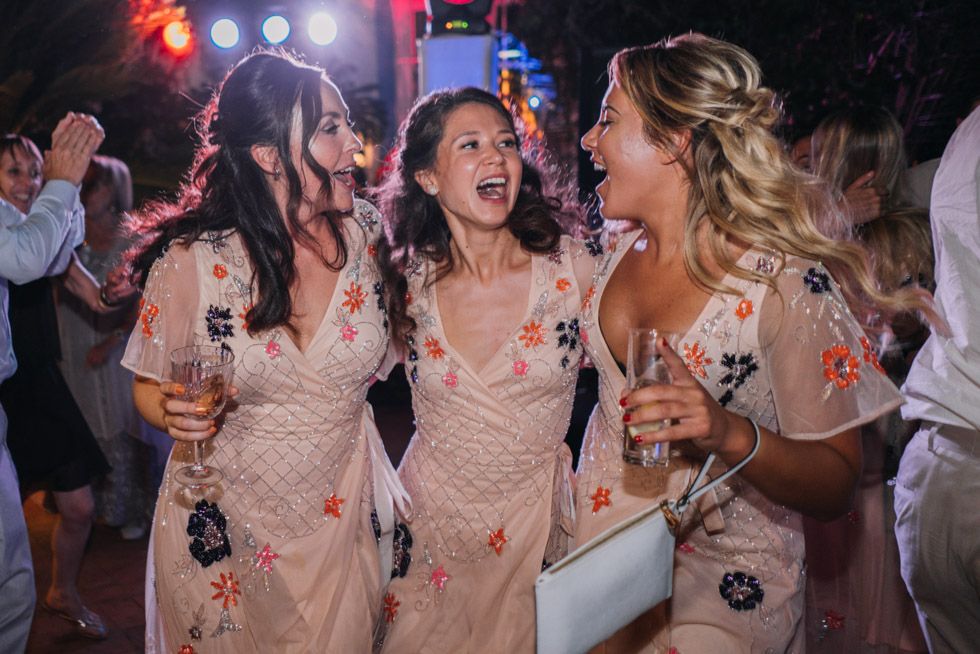 Three women in pink and floral dresses sharing joyful laughter during a celebration party with drinks in hand.