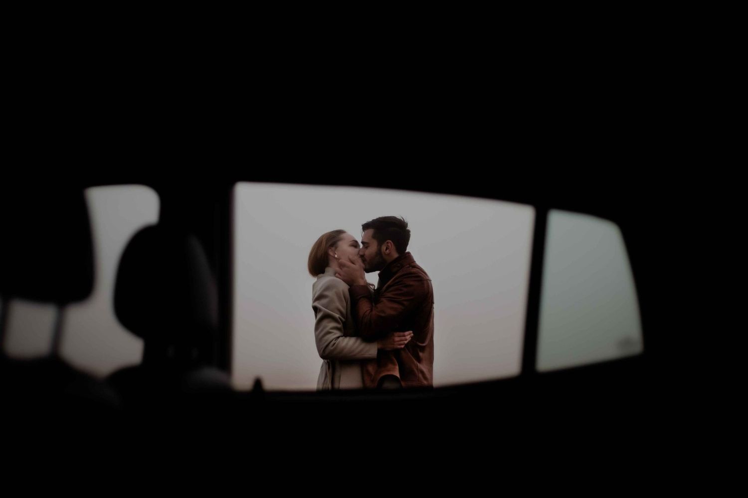 A romantic silhouette viewed through a car mirror shows a couple embracing at sunset.