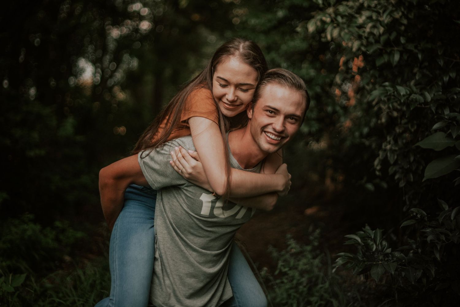 A playful piggyback ride moment captured between a joyful couple in a lush forest setting.