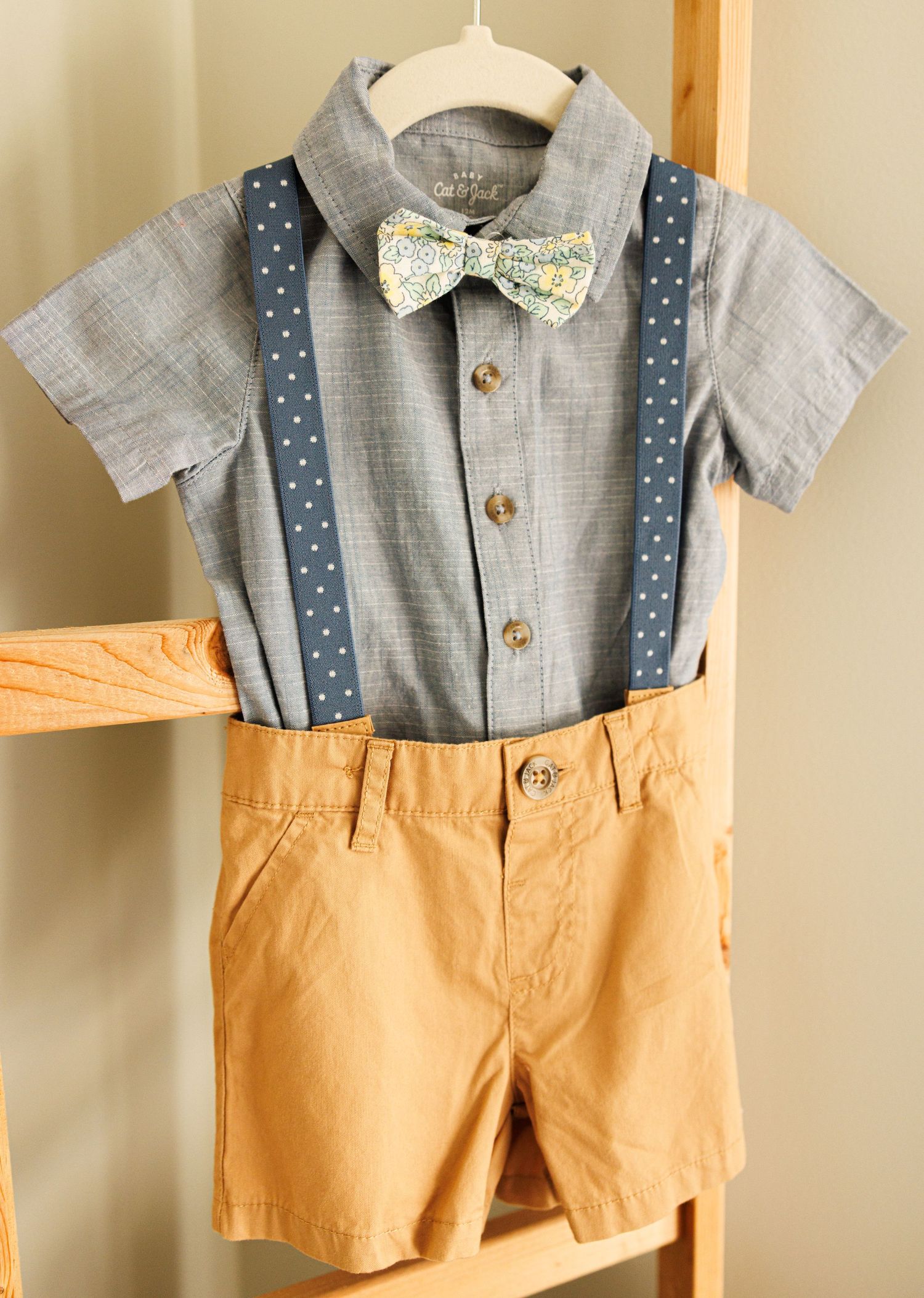 Stylish kids outfit featuring gray button-down shirt with floral bow tie, navy suspenders and khaki shorts.