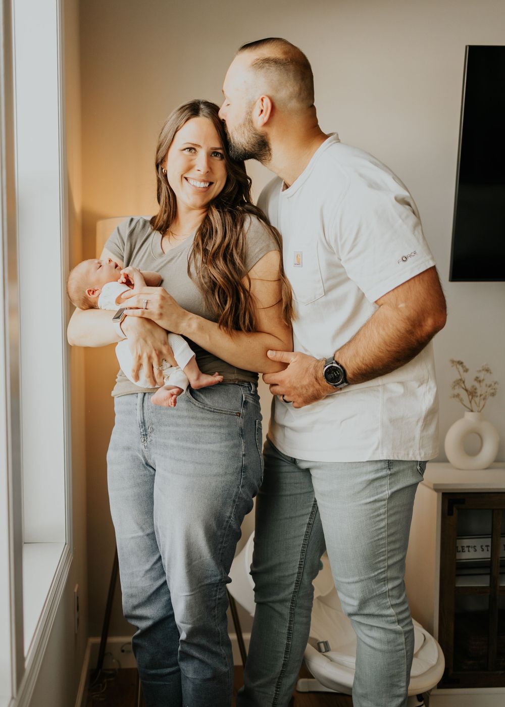 A couple in casual clothing stands near a window sharing an intimate moment while holding a newborn baby in warm lighting.