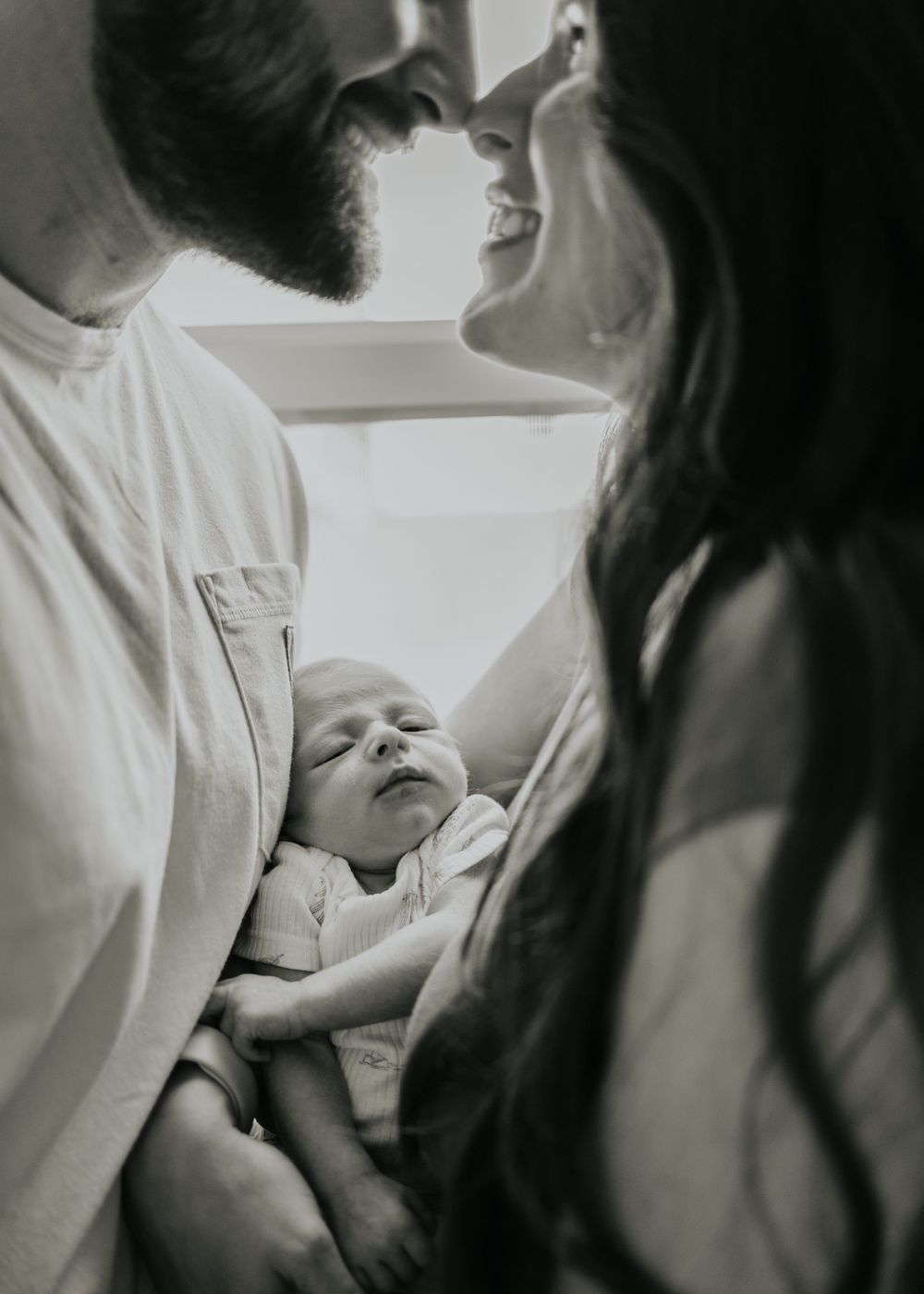 A tender black and white photo shows parents gazing lovingly at their sleeping newborn baby.