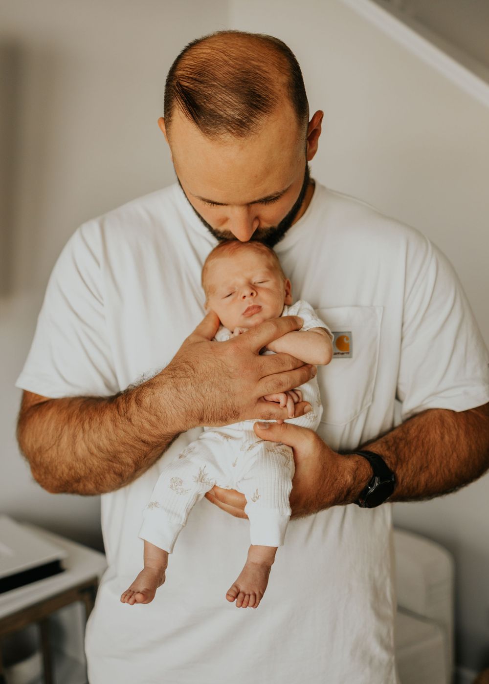 A tender moment captured as a parent in a white shirt cradles and kisses a sleeping newborn baby in a bright room.