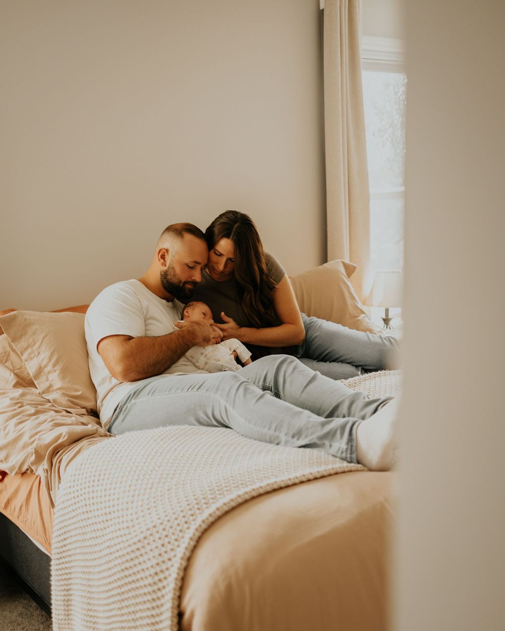 Couple relaxing together on a cozy bed with white linens near a window in warm natural lighting.