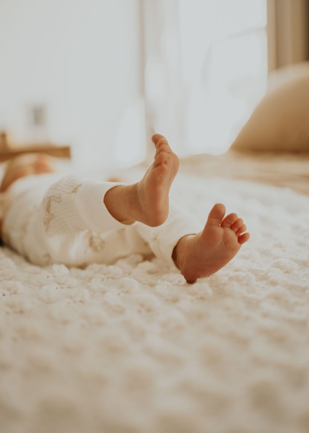 Small bare feet of a baby resting on a soft white textured carpet in warm natural lighting.
