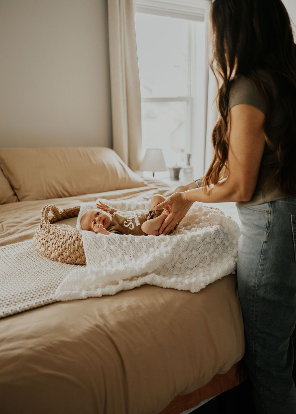 Person sitting on bed with knitted blanket and woven basket in warm sunlit bedroom with neutral tones.