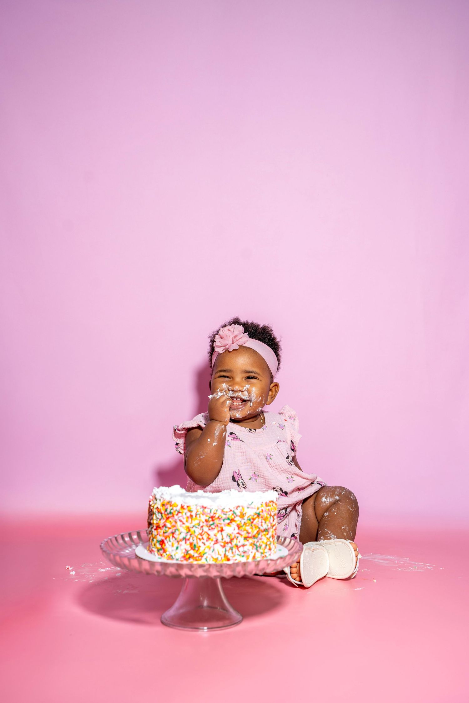 A small child sits on pink backdrop with a colorful sprinkle-covered birthday cake, wearing a pink outfit and headband.