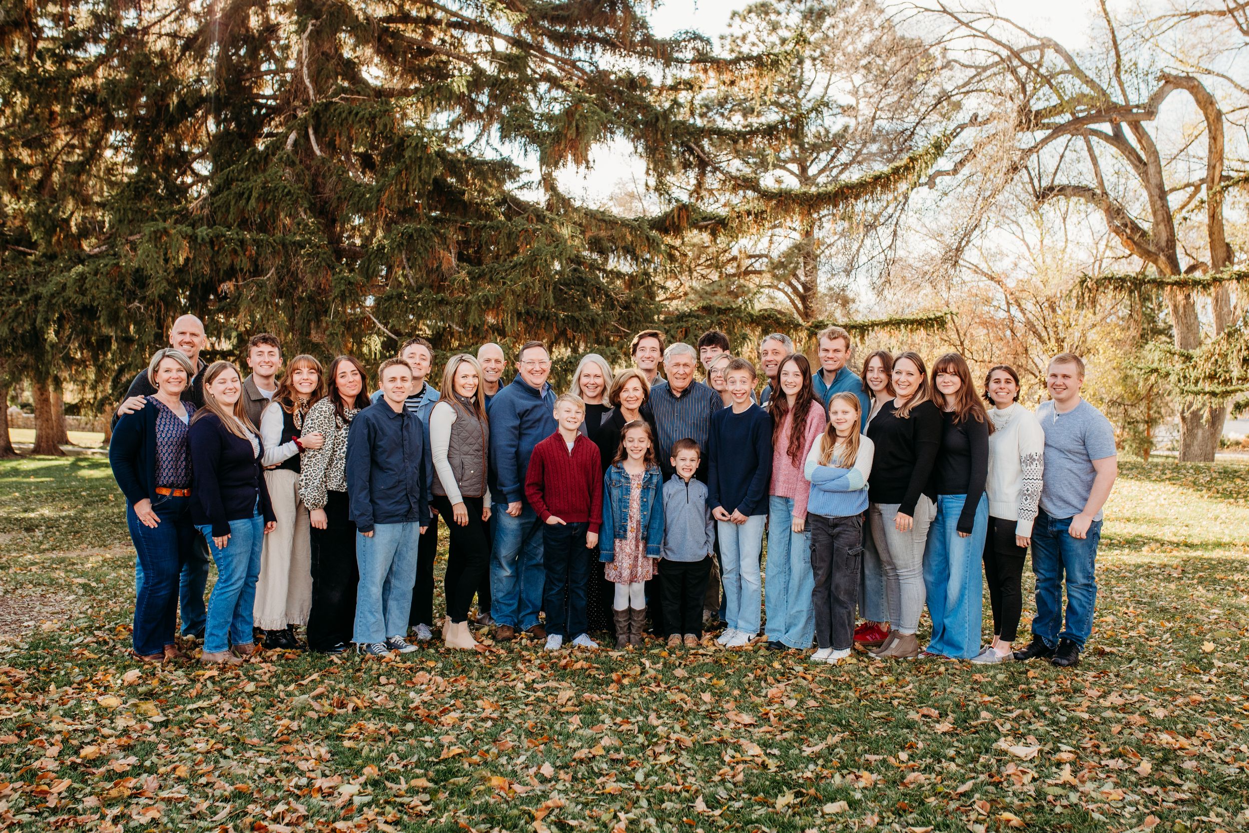 Large family group photo outdoors with fall trees and scattered leaves in background.