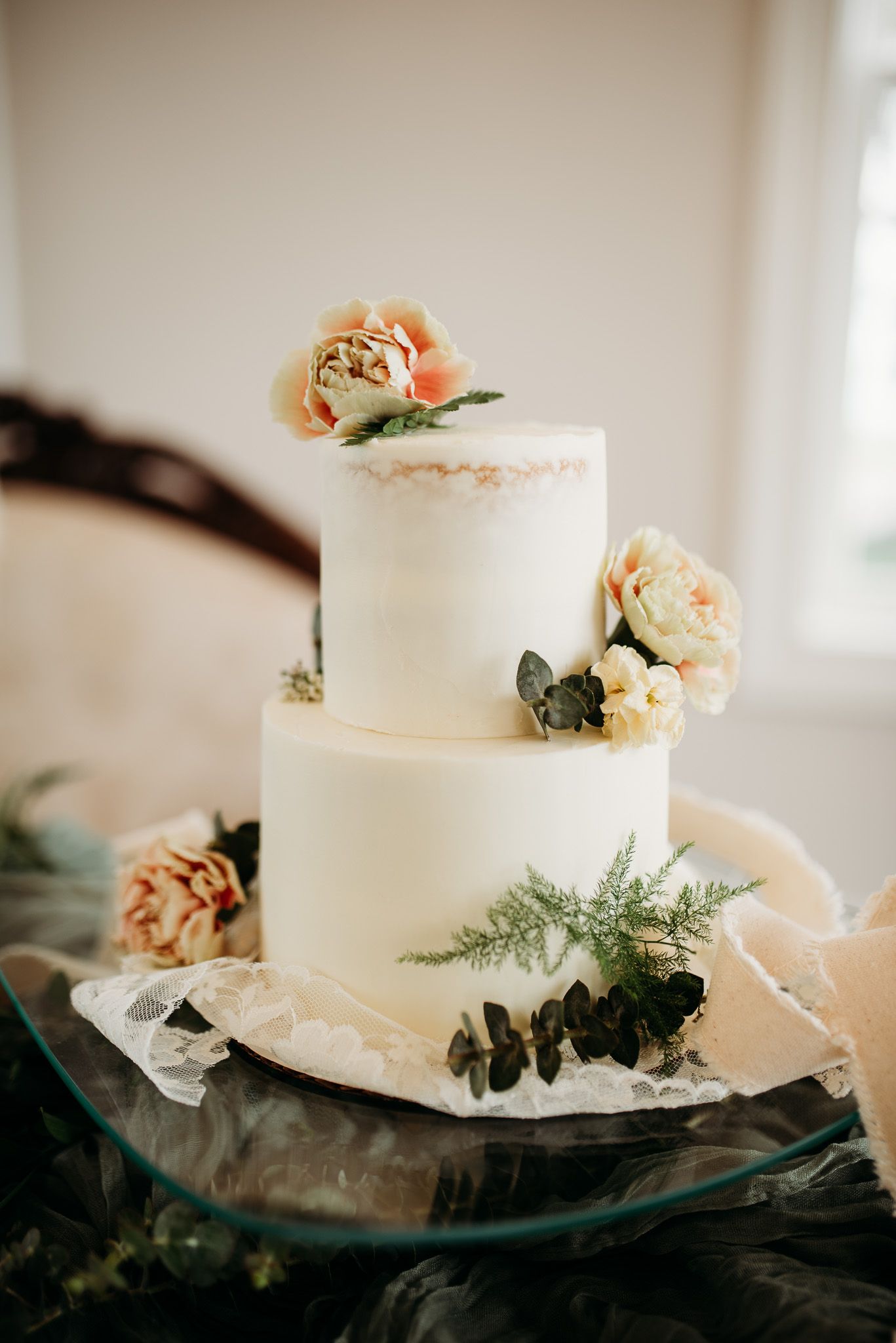 Elegant two-tier white wedding cake decorated with peach roses and greenery on a glass stand.