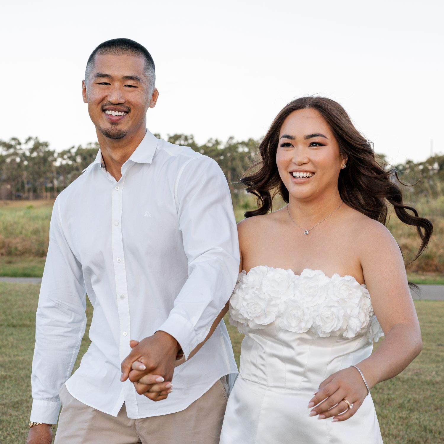 A couple in white formal attire poses on a grassy field during sunset.