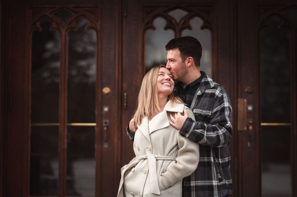 A couple embraces outdoors while wearing winter coats in front of a wooden door with Gothic architectural details.