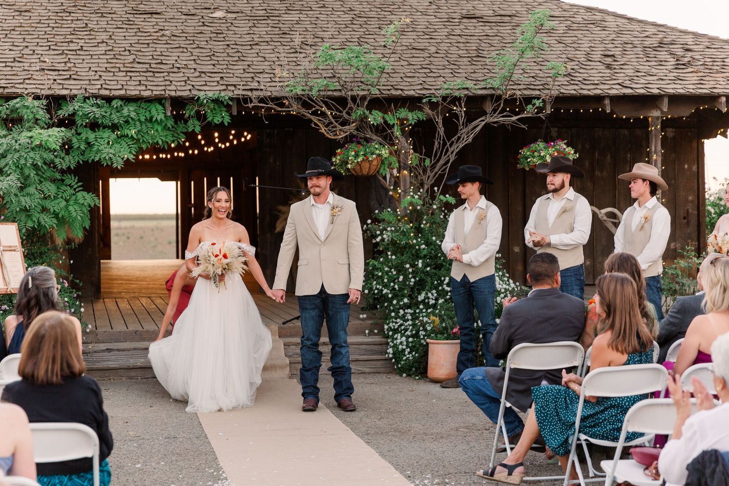 Wedding reception guests gather under string lights at an outdoor rustic venue for an evening celebration.