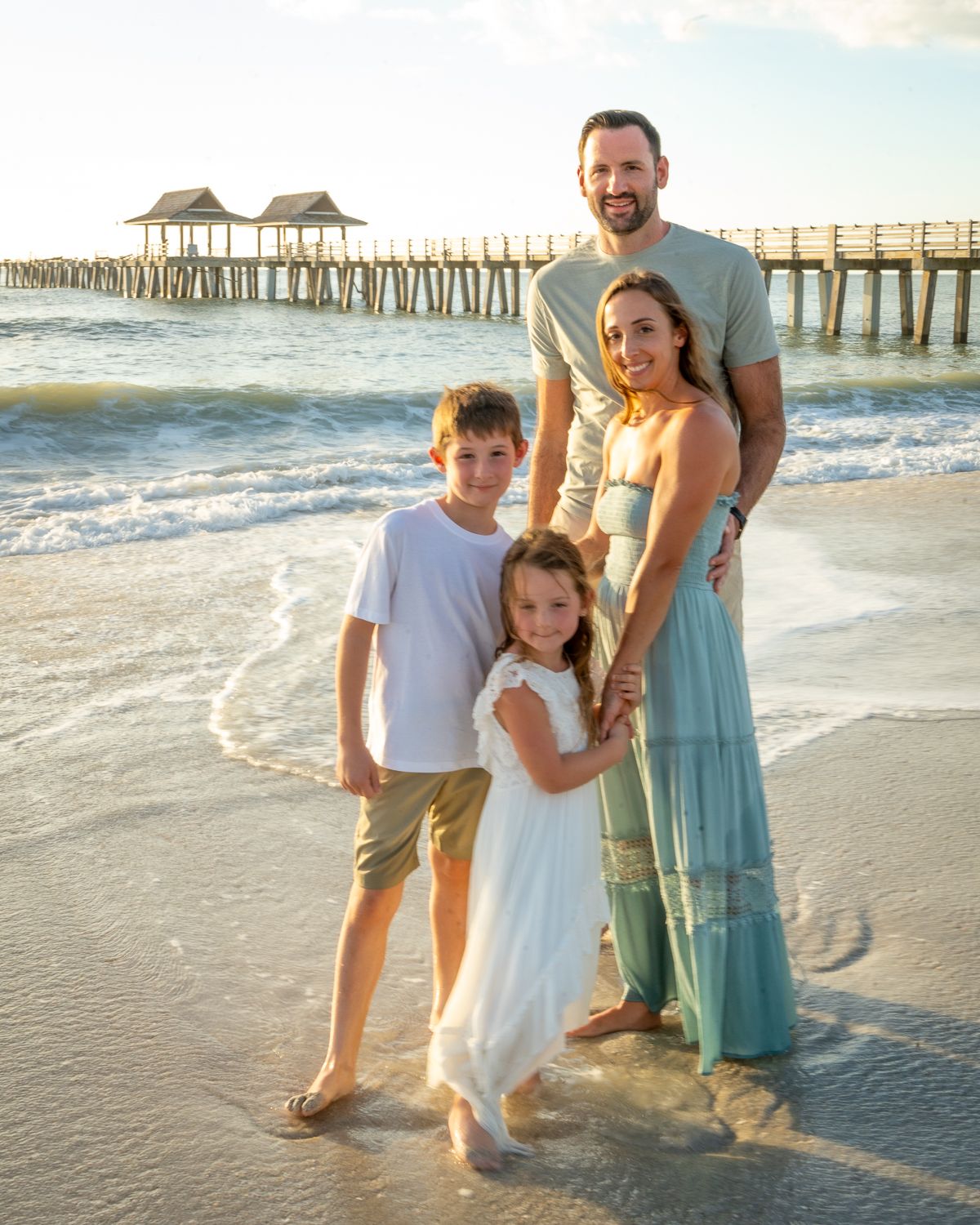 Family beach portraits near a fishing pier with coordinated blue and white outfits at sunset.