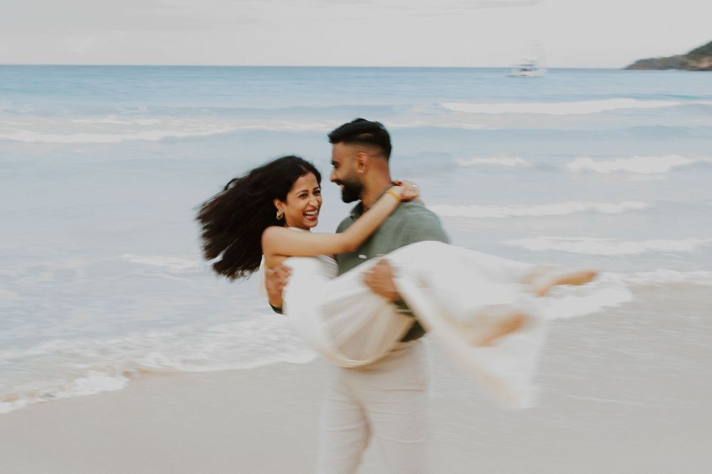Couple dancing and twirling on white sand beach with turquoise ocean in background.