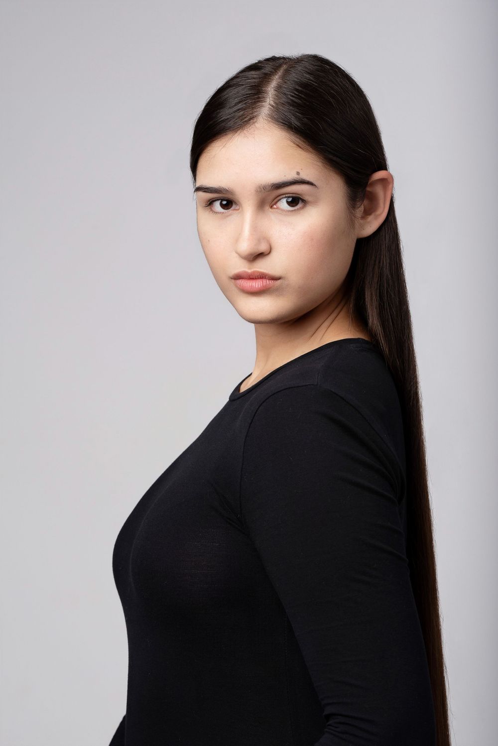 Studio portrait of a young woman with long dark hair wearing a black long-sleeved top against a light gray background.