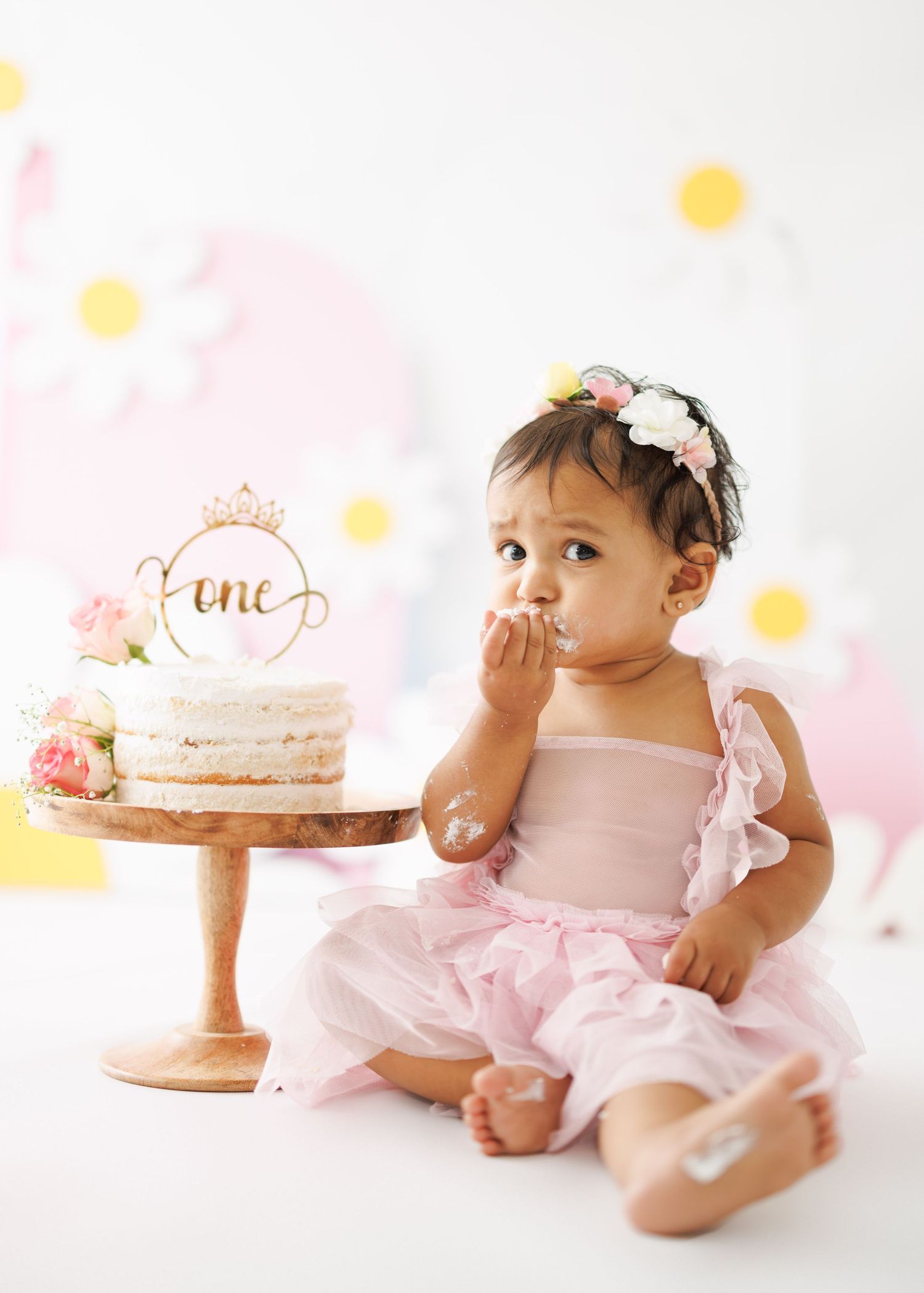 A first birthday celebration with a baby in pink dress sitting beside a decorated cake with gold accents.