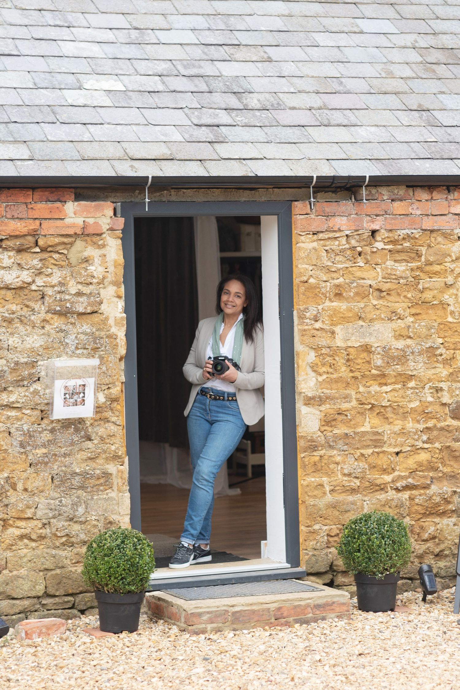Person standing in stone cottage doorway with vintage camera, flanked by boxwood plants and rustic stone walls.