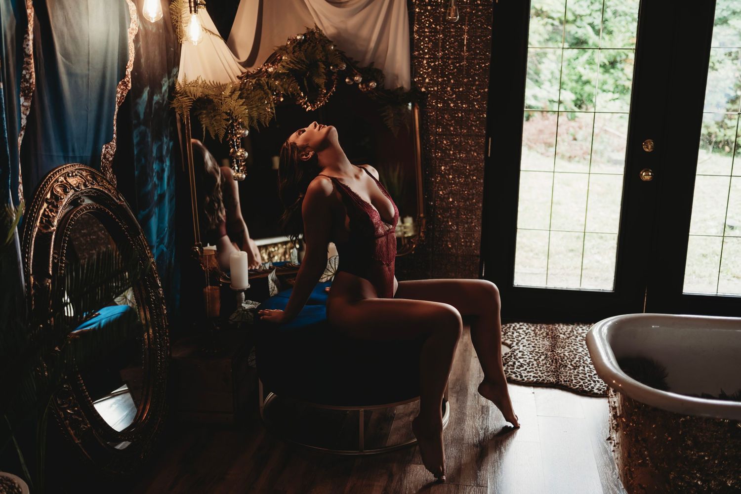 Woman in red lingerie, seated with an arched back in a cozy, vintage-inspired space, with a large mirror and a clawfoot tub.