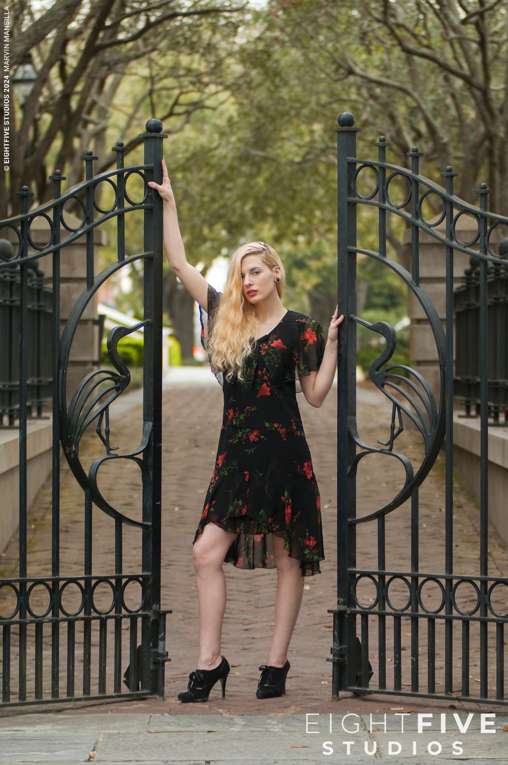 Blonde woman in black floral dress and heels posing at ornate iron gate outdoors.