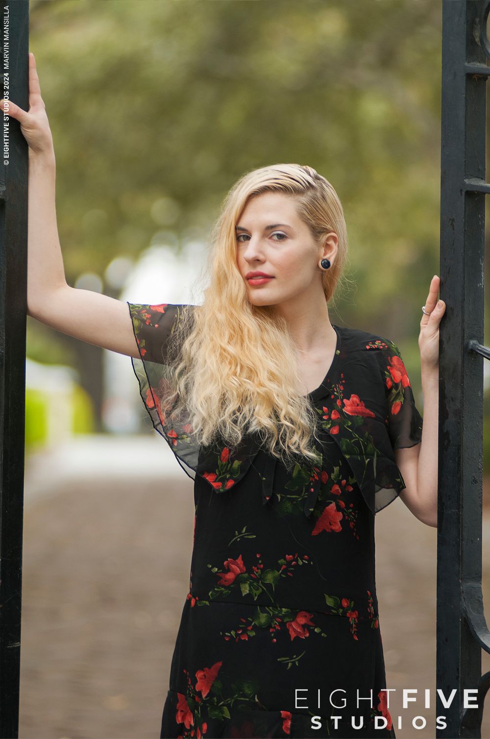 Blonde woman in black floral dress posing at iron gate by Eight Five Studios.