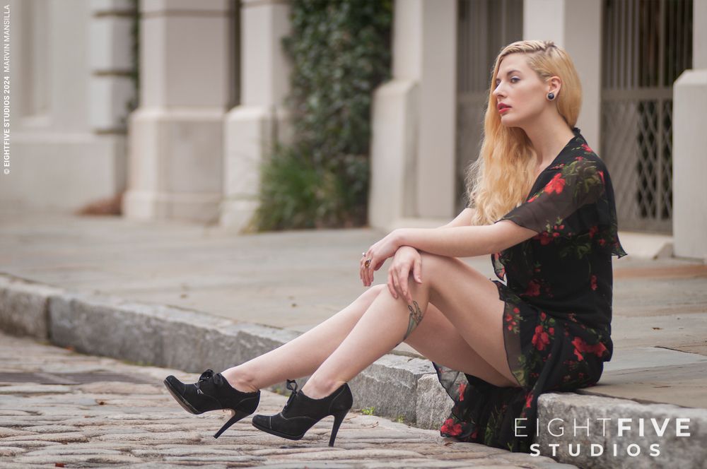 Blonde woman in black floral dress and heels sitting on urban steps outdoors.