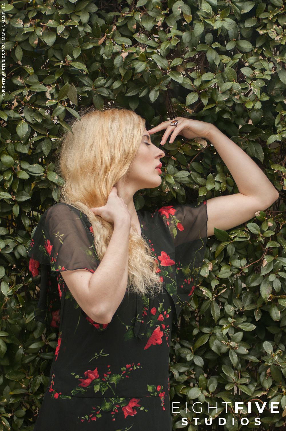 Blonde woman in black floral dress posing against lush green hedge wall.