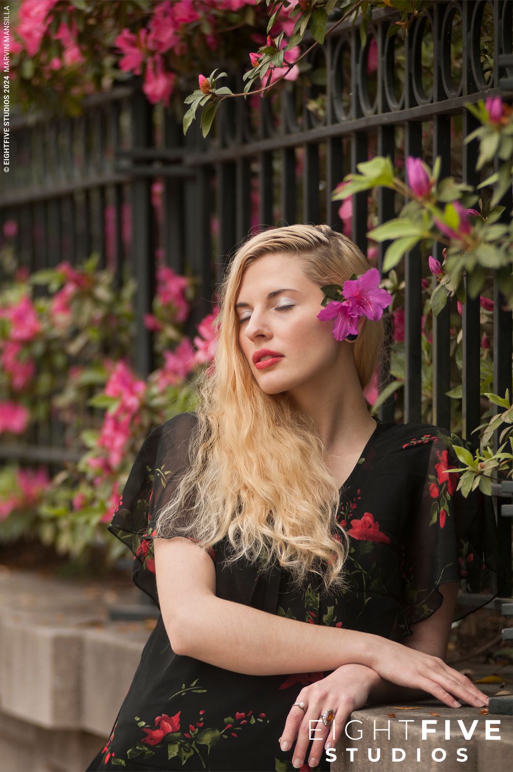 Blonde woman with pink flower in hair posing by iron fence with bougainvillea.