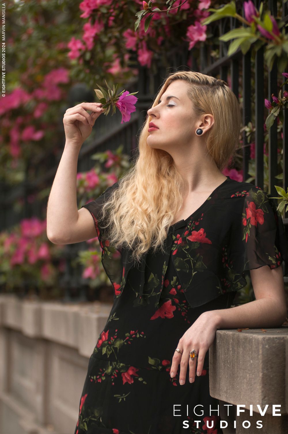 Blonde woman smelling pink flower near iron fence with bougainvillea blooms.