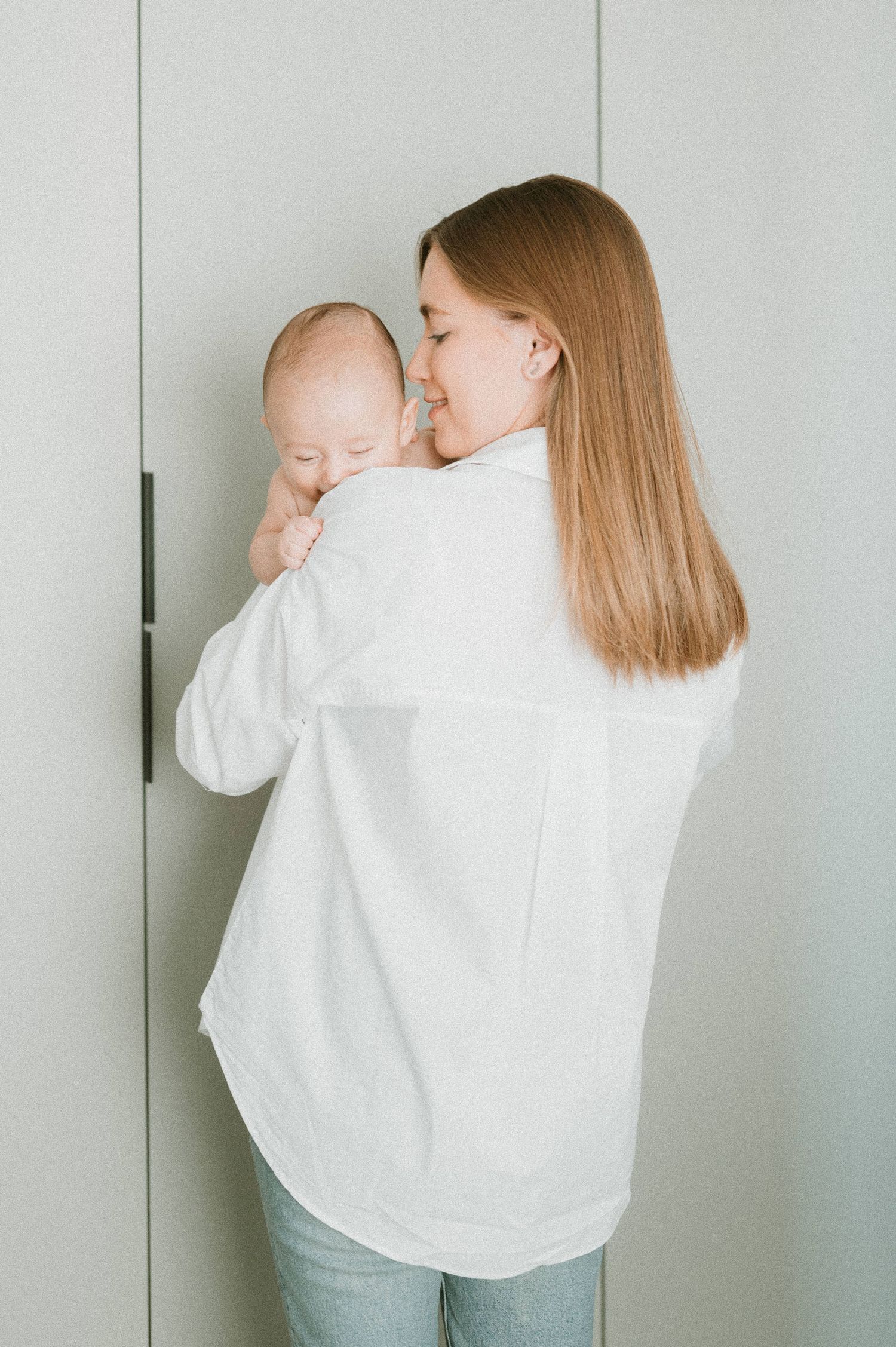 Parent in white shirt holding and comforting infant against neutral wall background.