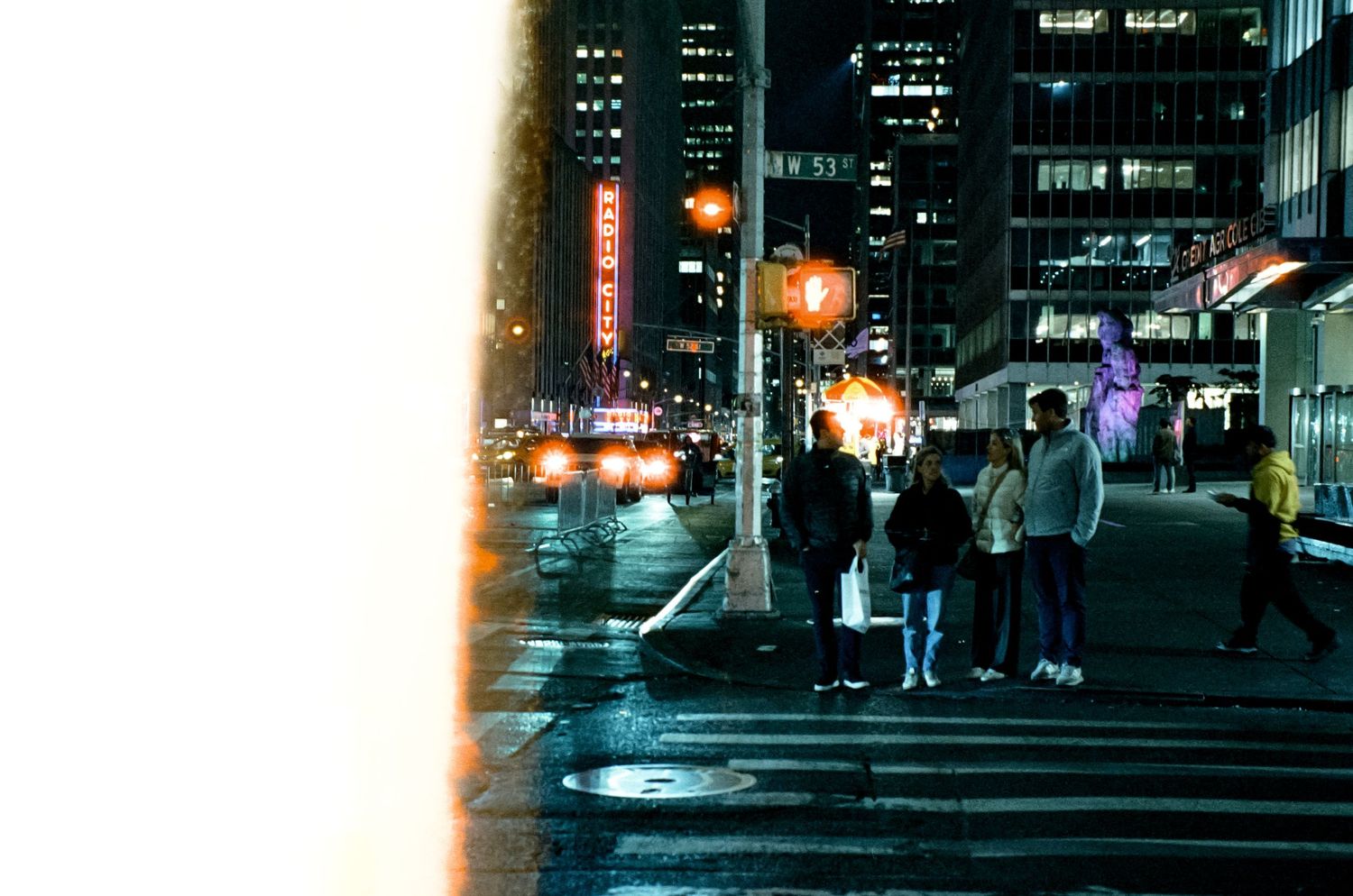 Nighttime city street scene with illuminated buildings and people waiting at crosswalk.
