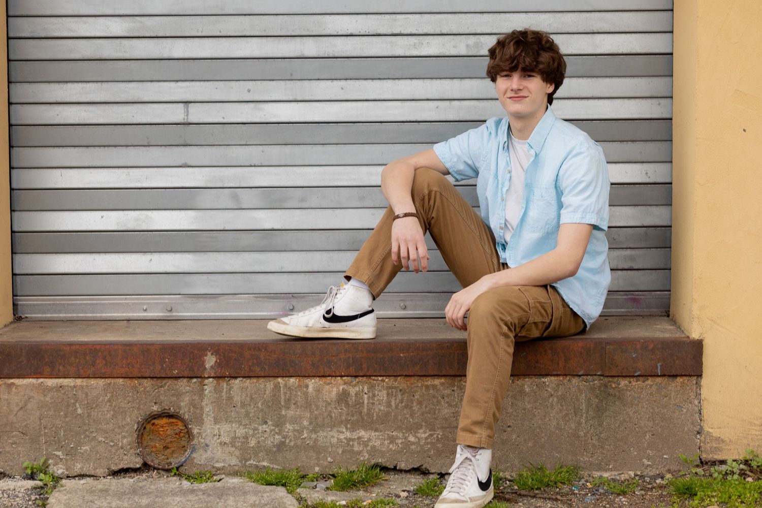Casual portrait of a teen sitting on a concrete ledge against industrial metal shutters.
