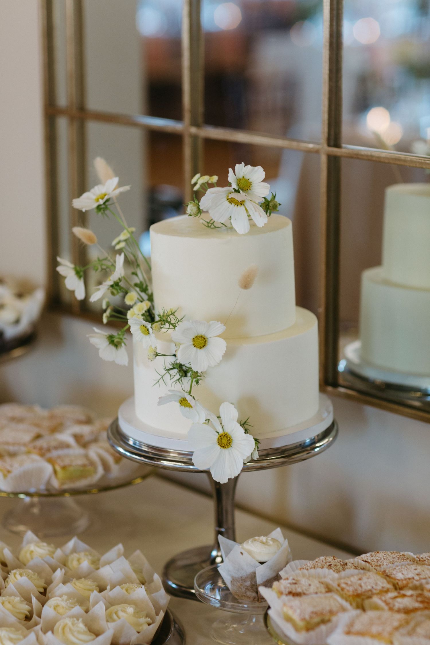 A two-tiered white wedding cake decorated with delicate white daisies on a glass pedestal stand.
