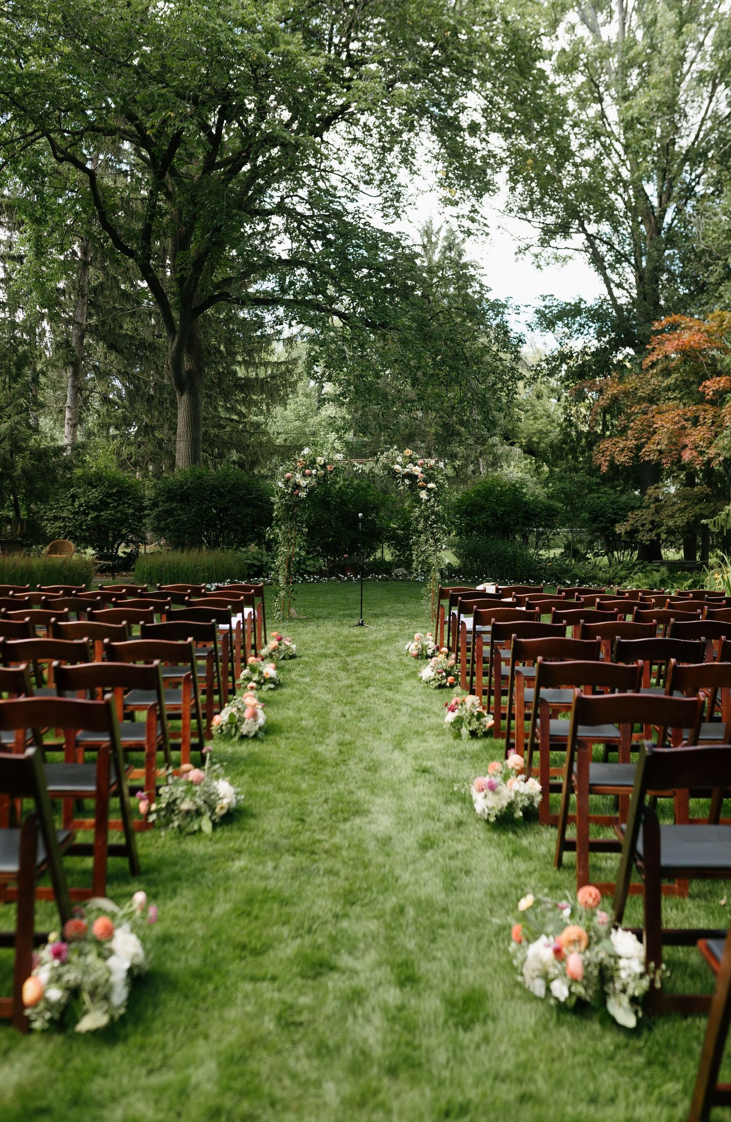 Outdoor wedding ceremony setup with wooden chairs and floral aisle decorations in a lush garden setting.