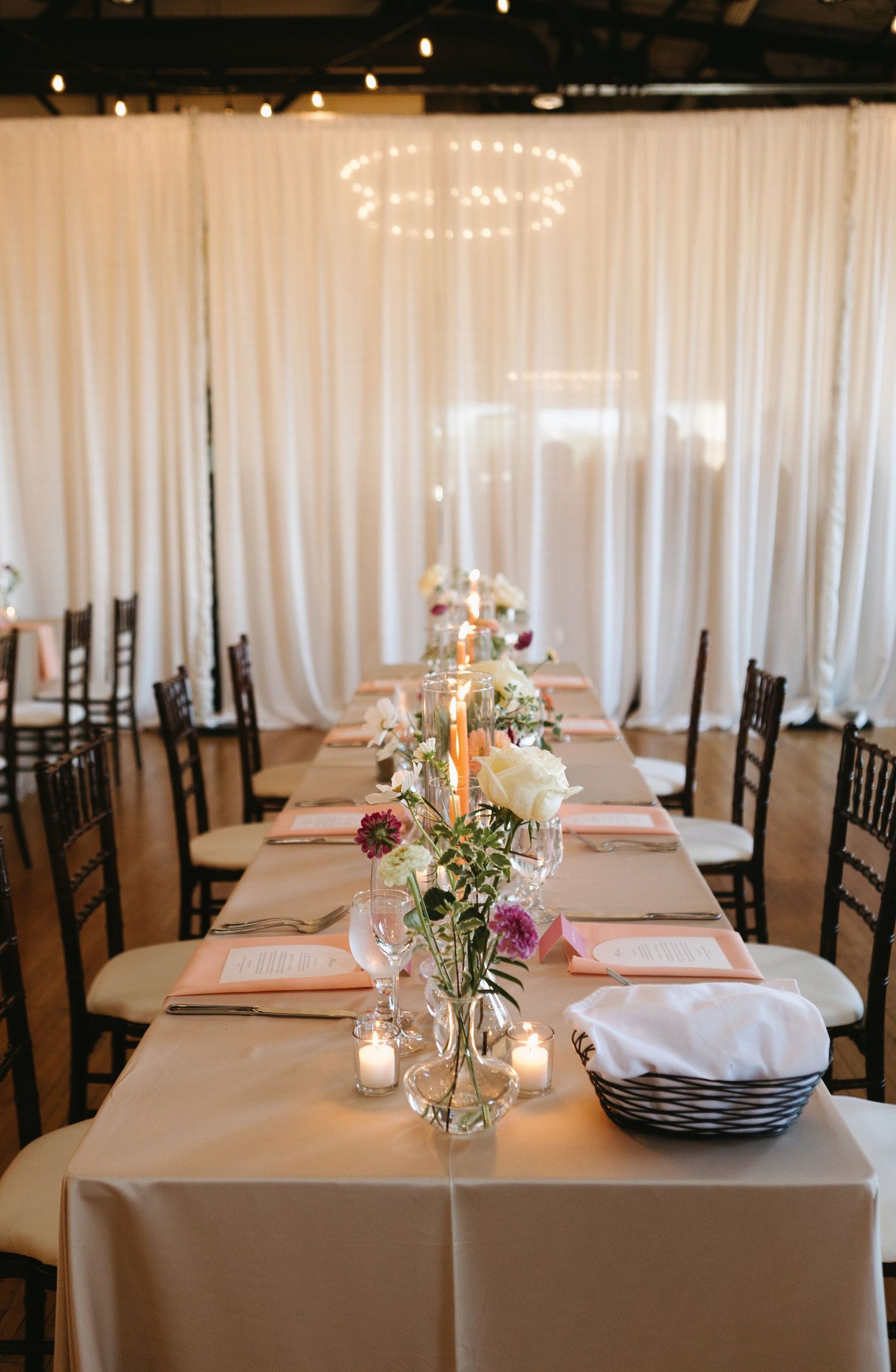 Long banquet table set with white linens, candlelight, and simple floral arrangements against white draping.