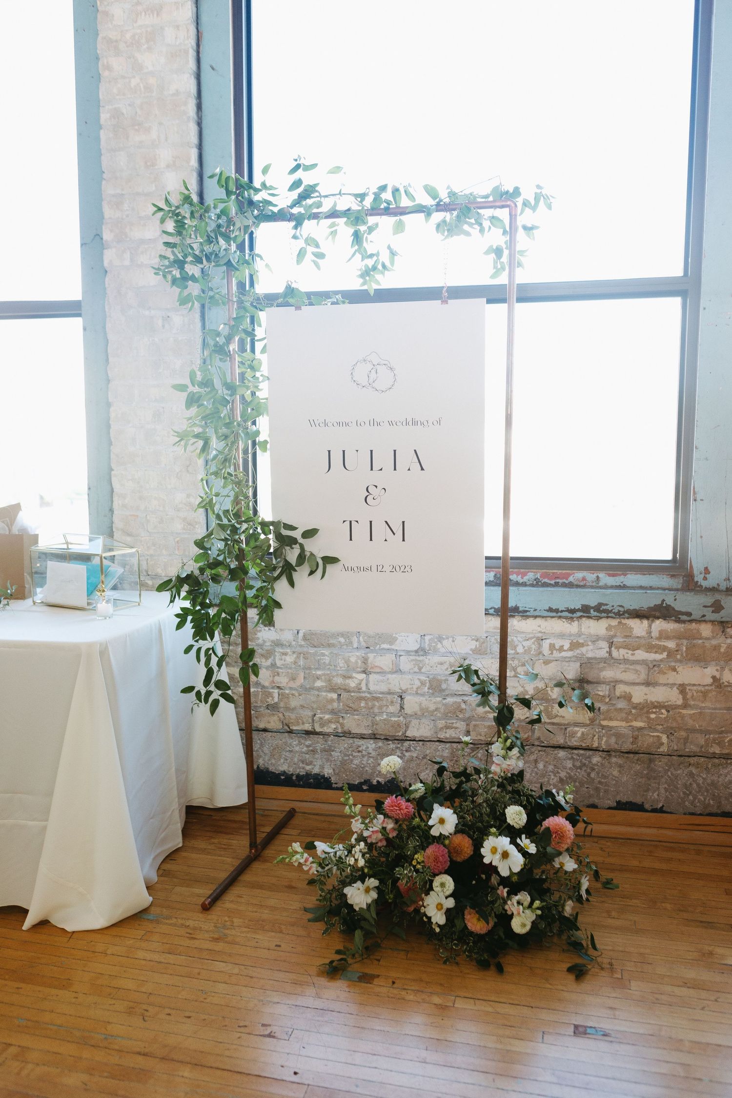 Wedding welcome sign with greenery and floral decorations against exposed brick wall.
