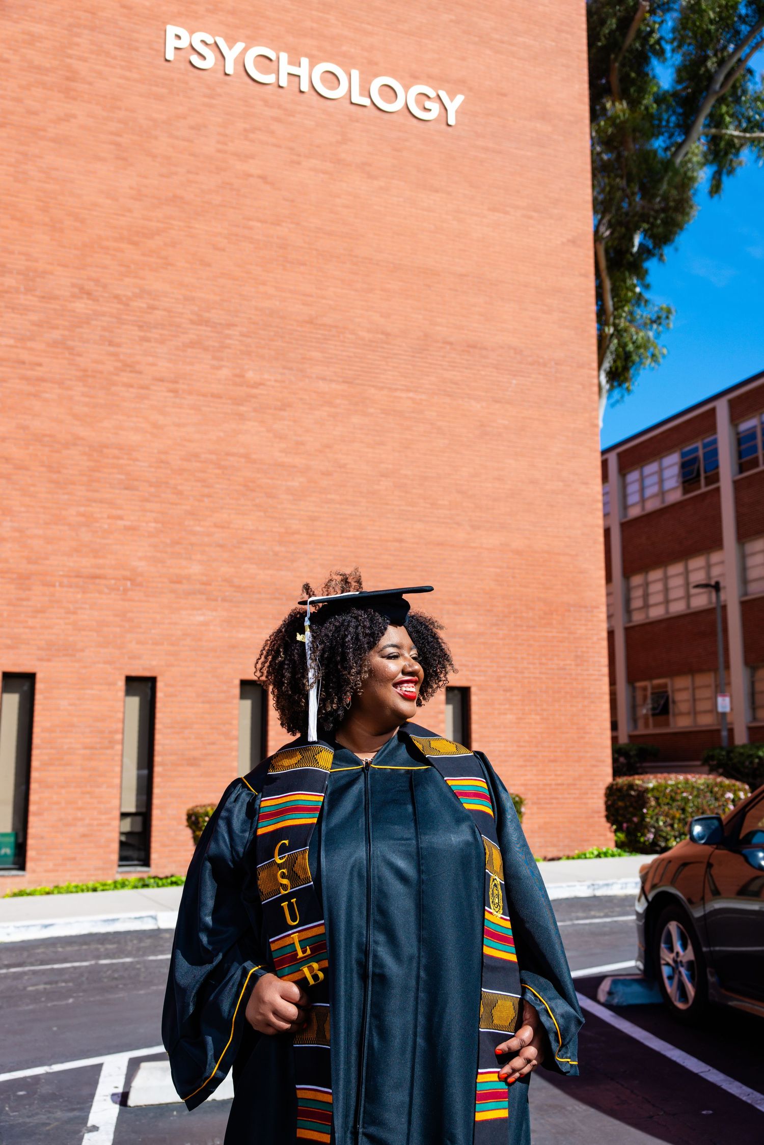 Graduate in cap and gown poses in front of Psychology building on sunny day.
