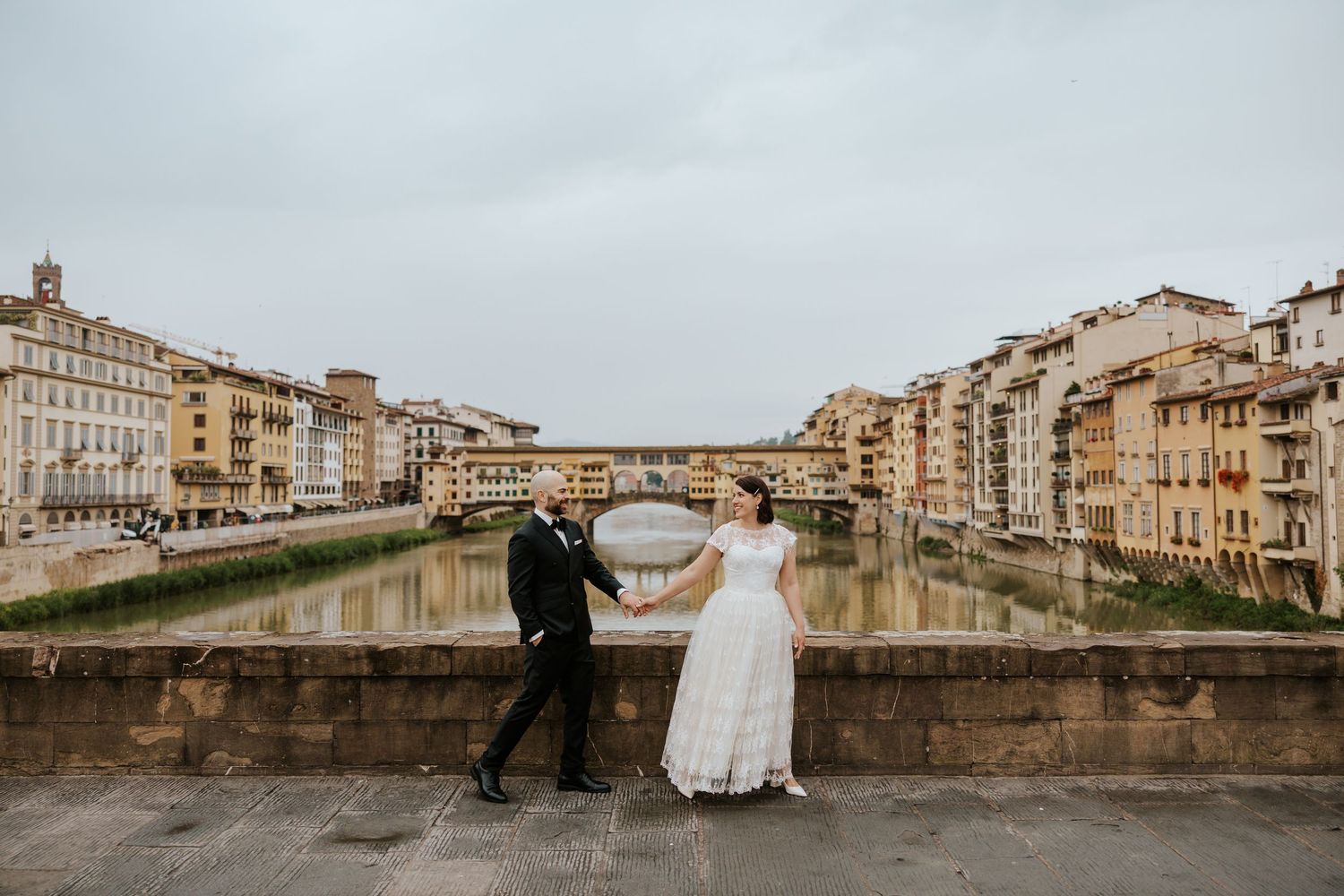 playful couple in florence italy walking across a bridge with a view of the ponte vecchio