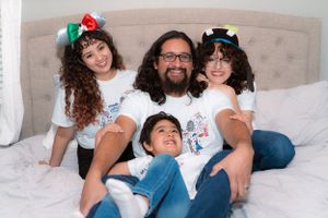 Family of four in matching white Disney shirts and colorful mouse ears sitting on a bed.