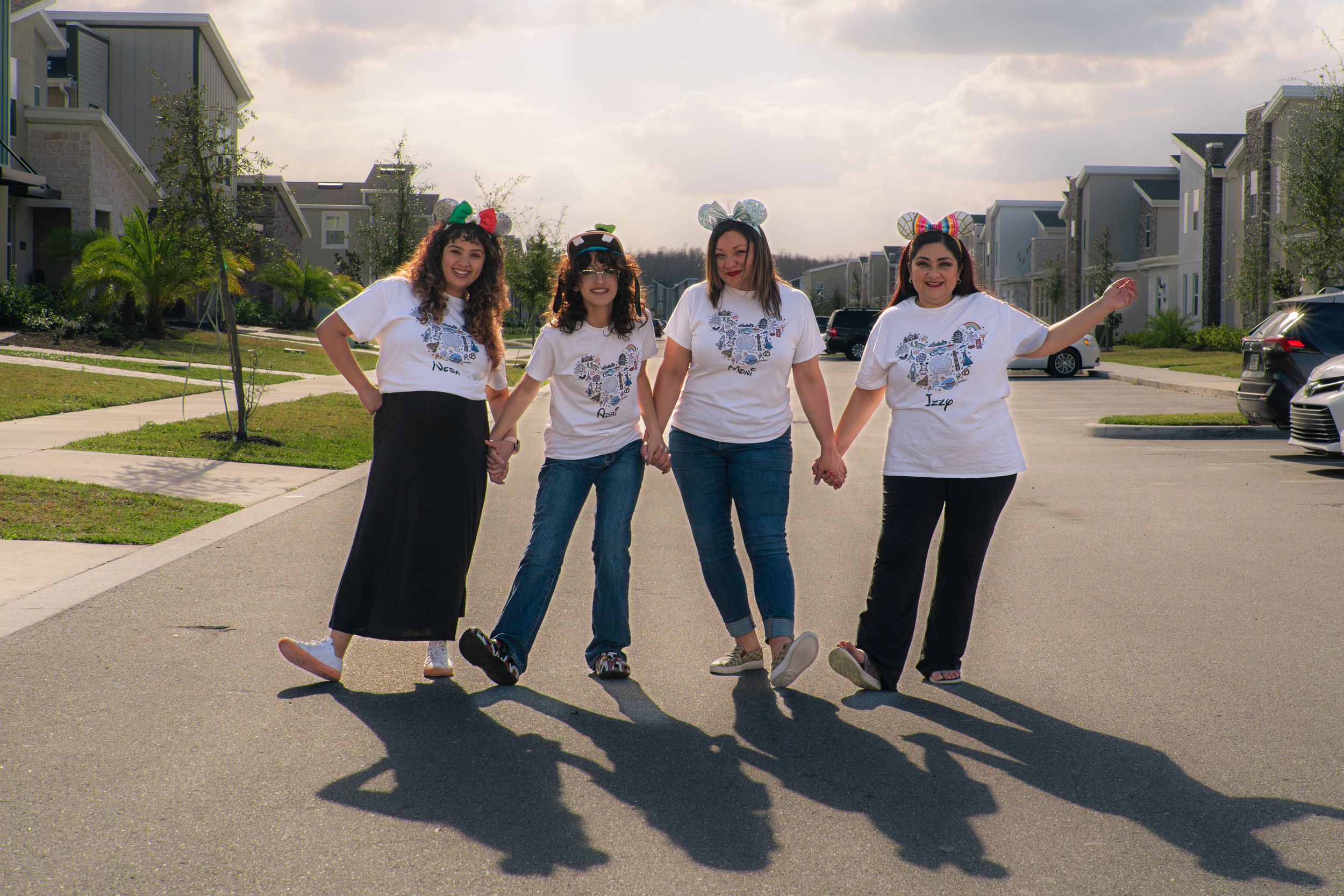 Four women in white shirts holding hands and posing on a sunny suburban street.