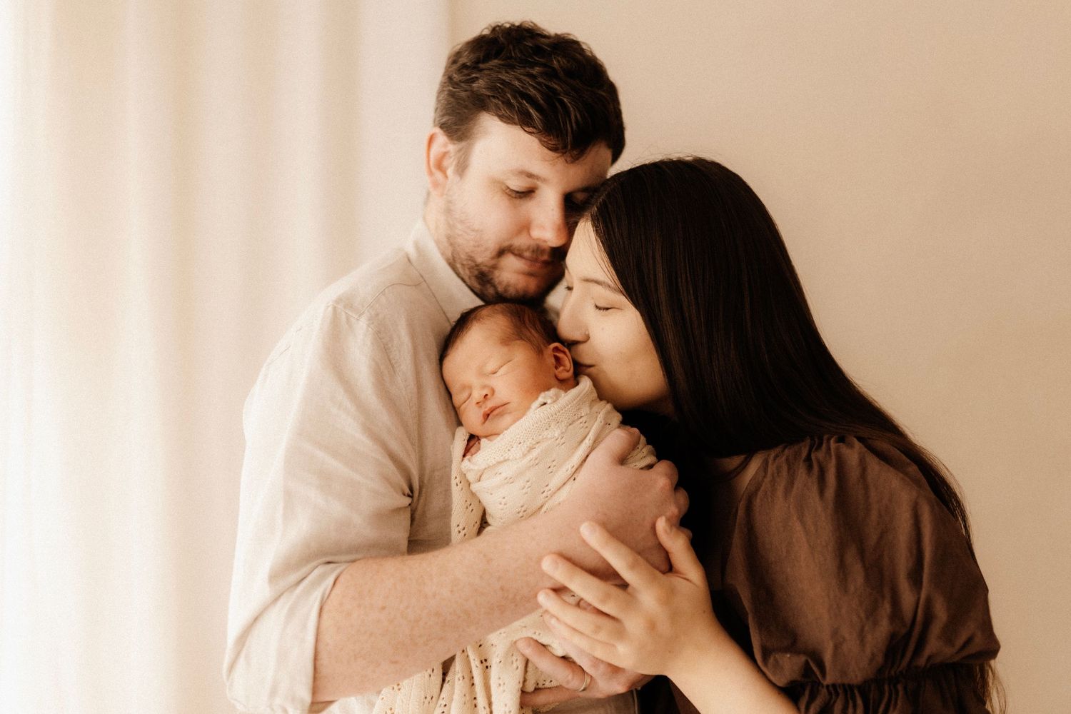 Tender family moment with newborn in warm neutral tones against white backdrop.