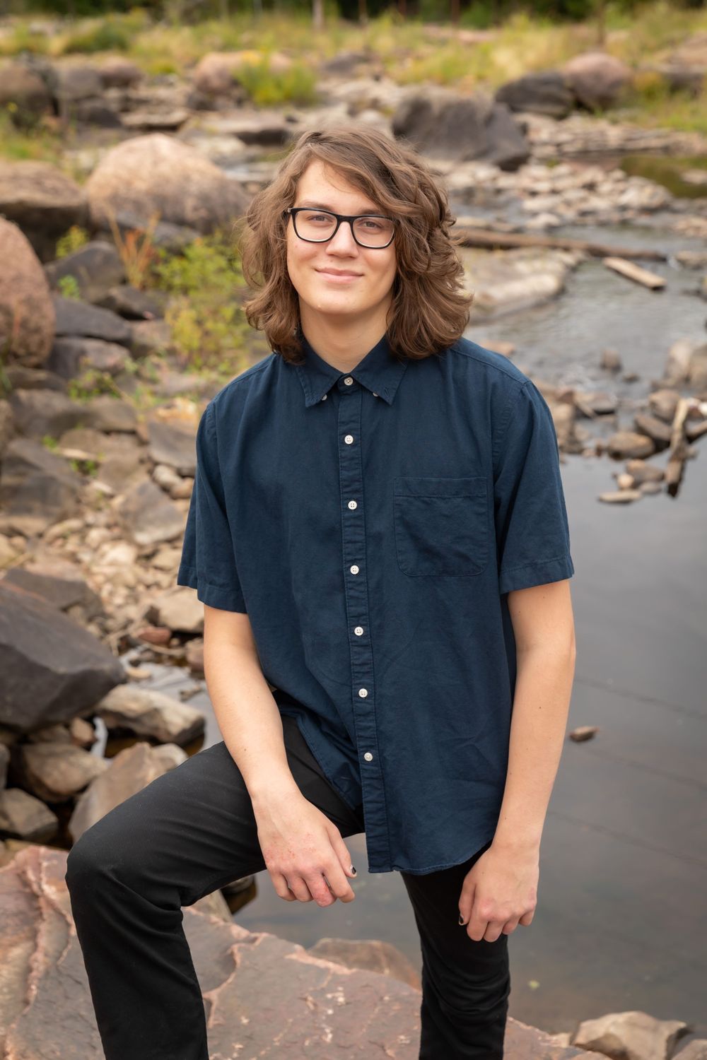 Casual outdoor portrait session on rocky terrain wearing navy button-down shirt and glasses.