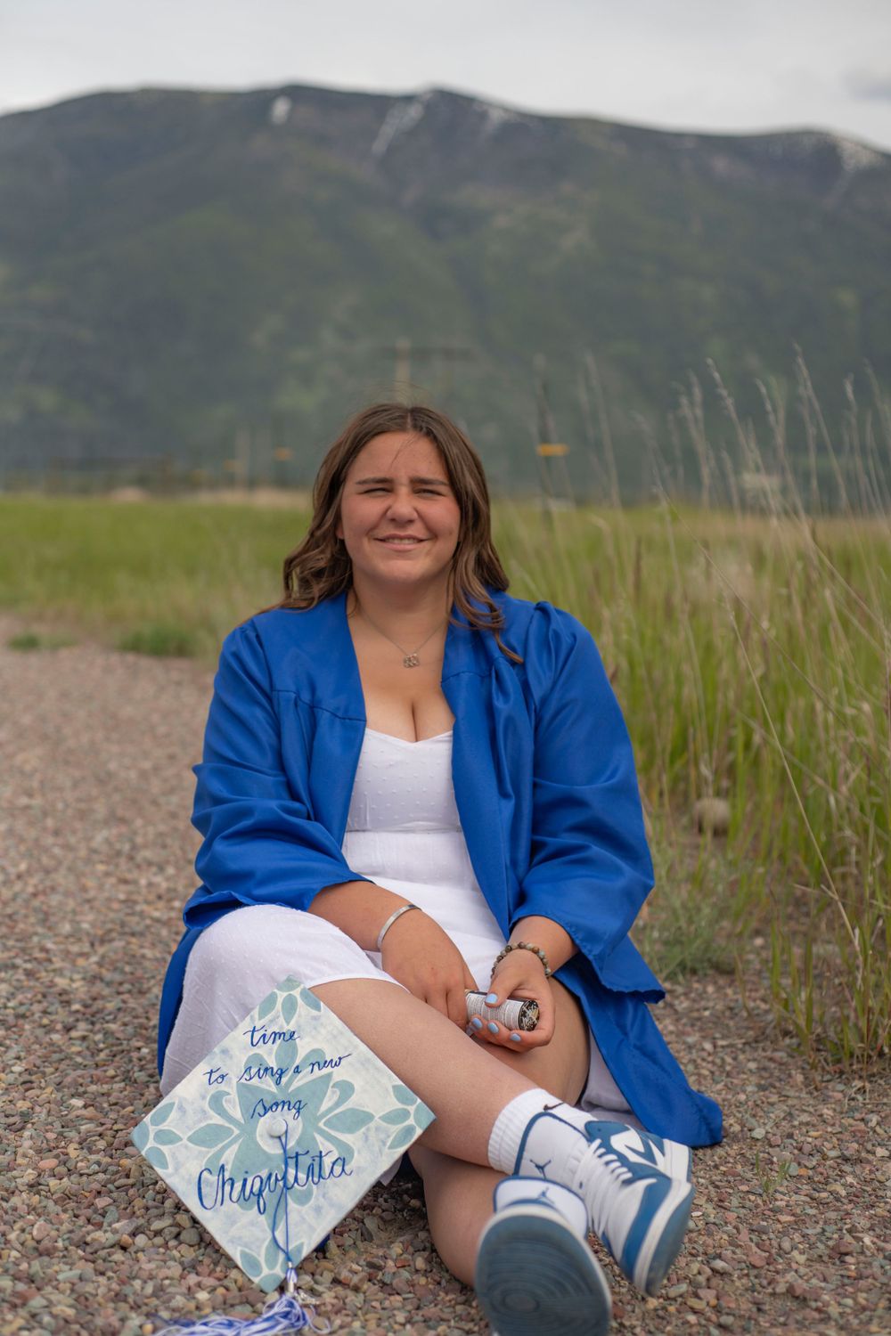 Young woman in blue graduation gown sits on gravel path with mountain backdrop holding decorated mortarboard.