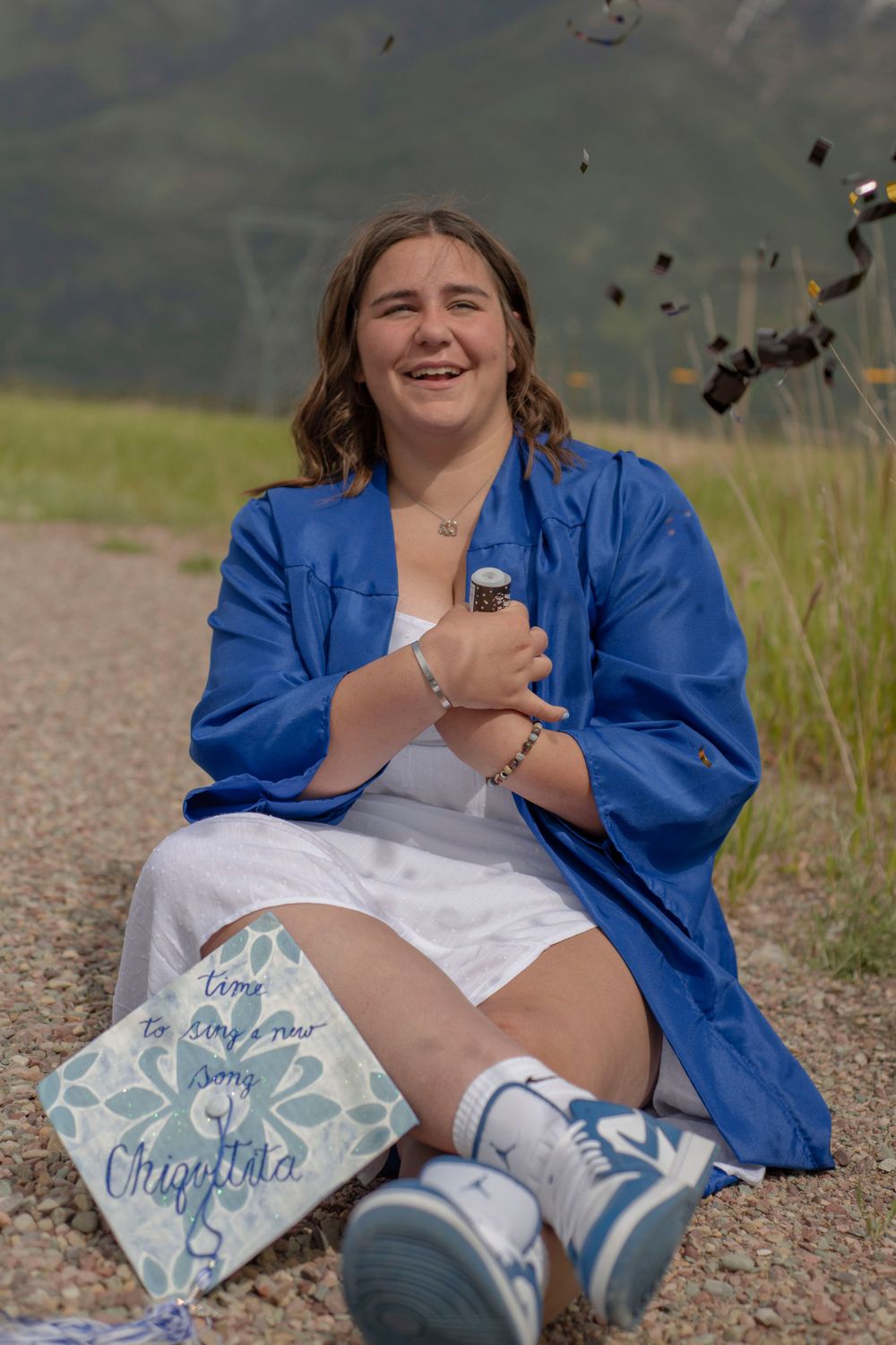 Joyful graduate in blue gown sitting on path with confetti falling.