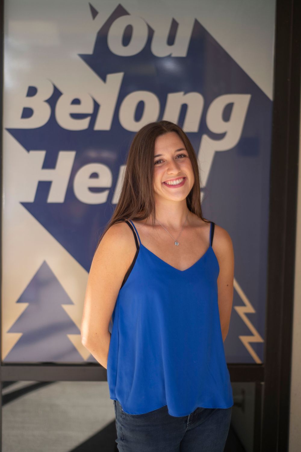 Smiling young woman in blue top stands before a You Belong Here sign.