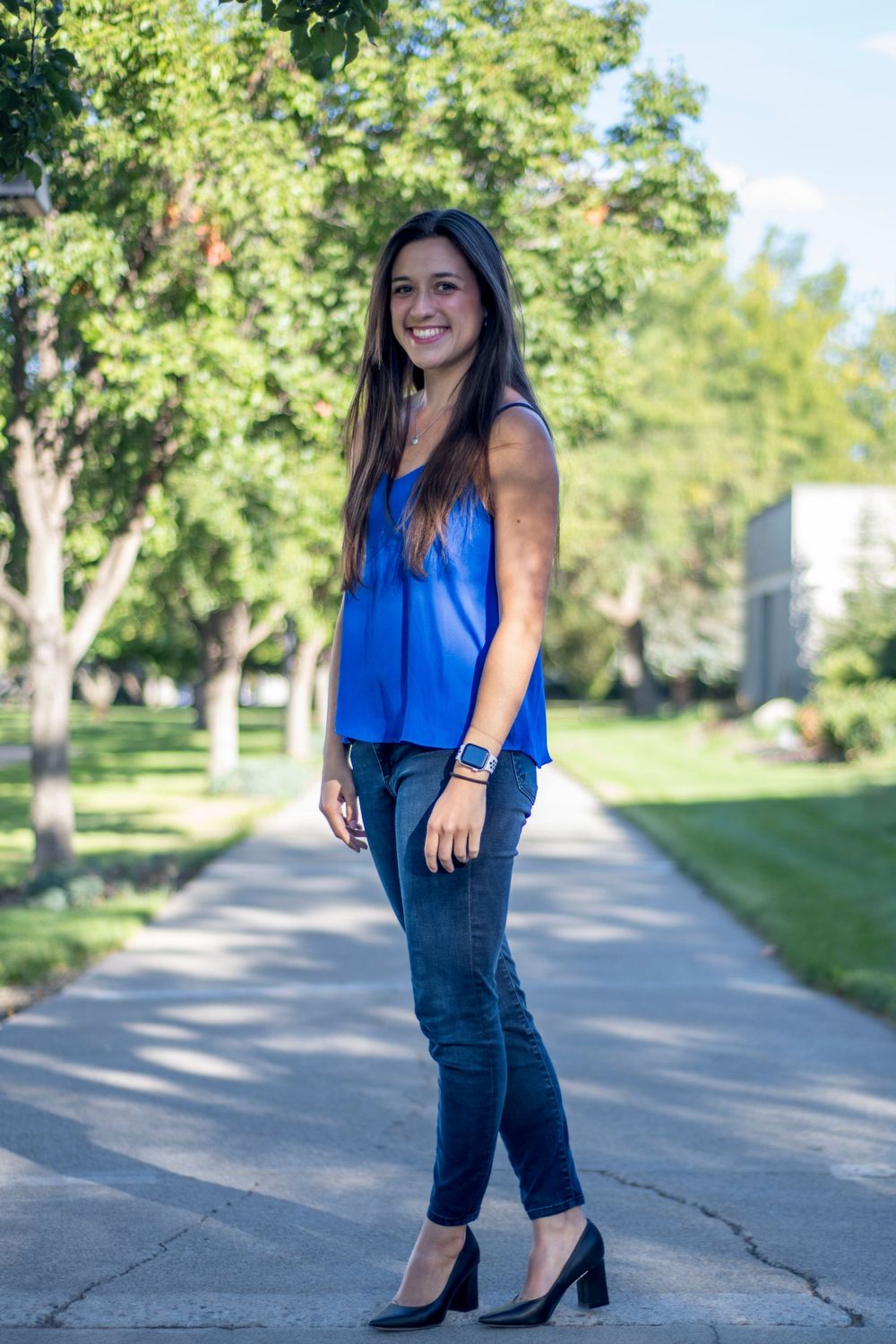 Smiling brunette woman in blue top and jeans poses on a tree-lined campus pathway.