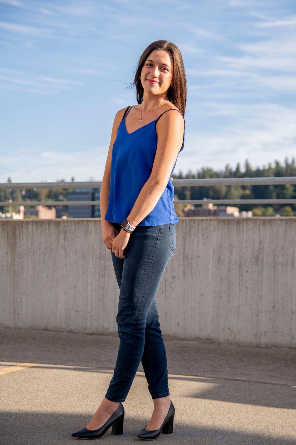 Young woman in royal blue camisole and dark jeans standing on rooftop under blue sky.